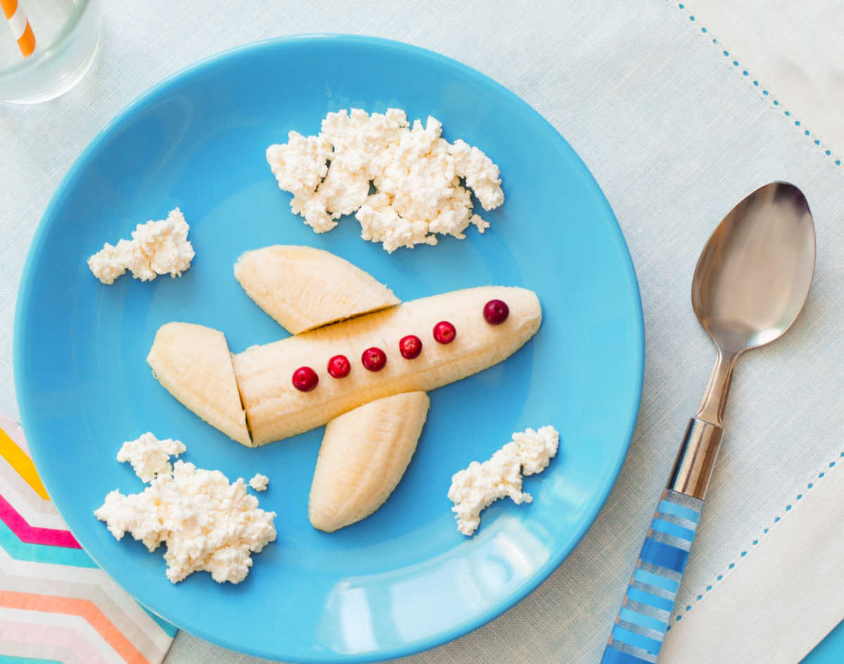 Banana food art - A banana in the shape of an airplane with some cottage cheese clouds, on a blue plate next to a spoon.