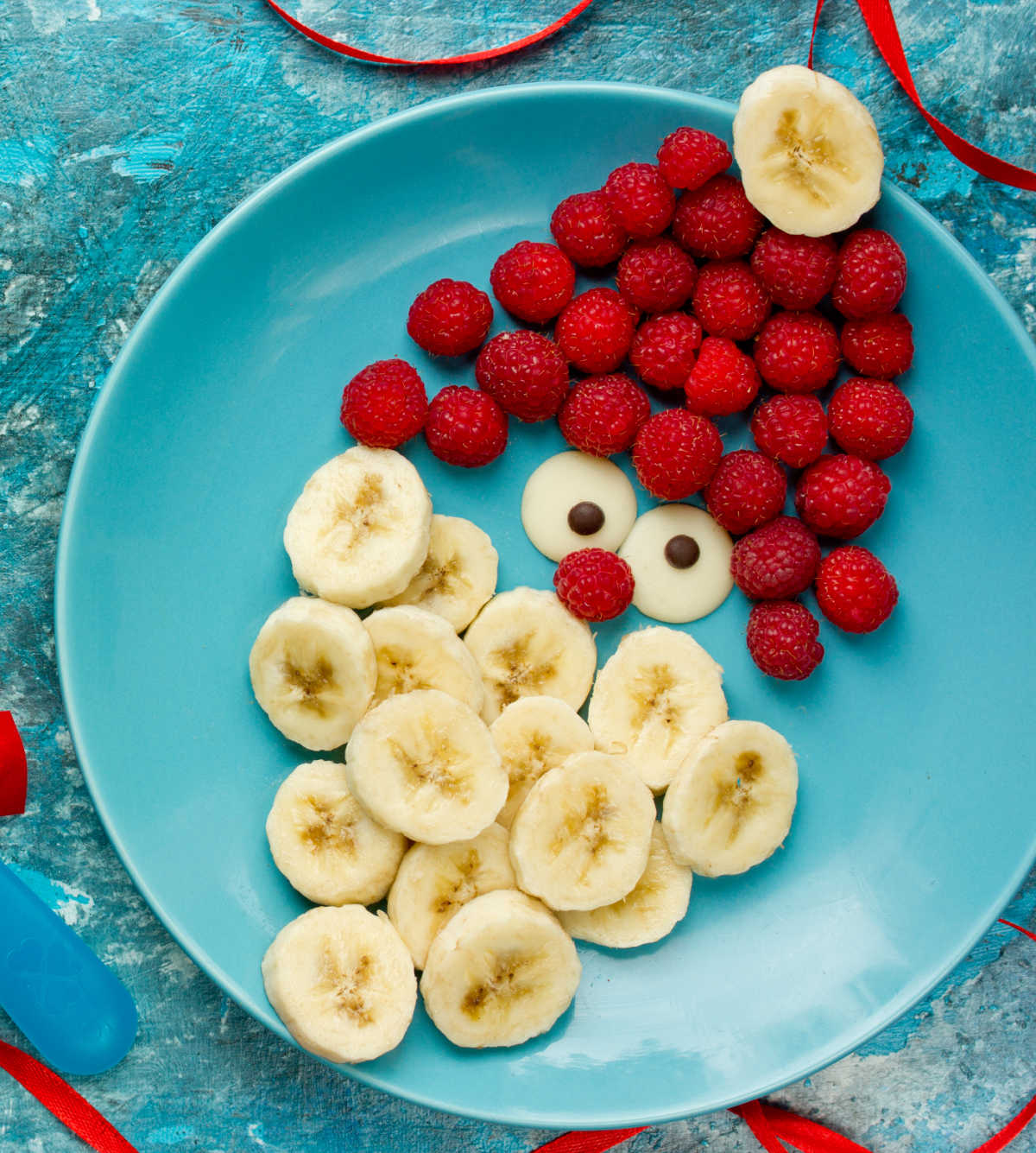 Raspberries and banana slices shaped to form a Santa Claus figure on a blue plate.