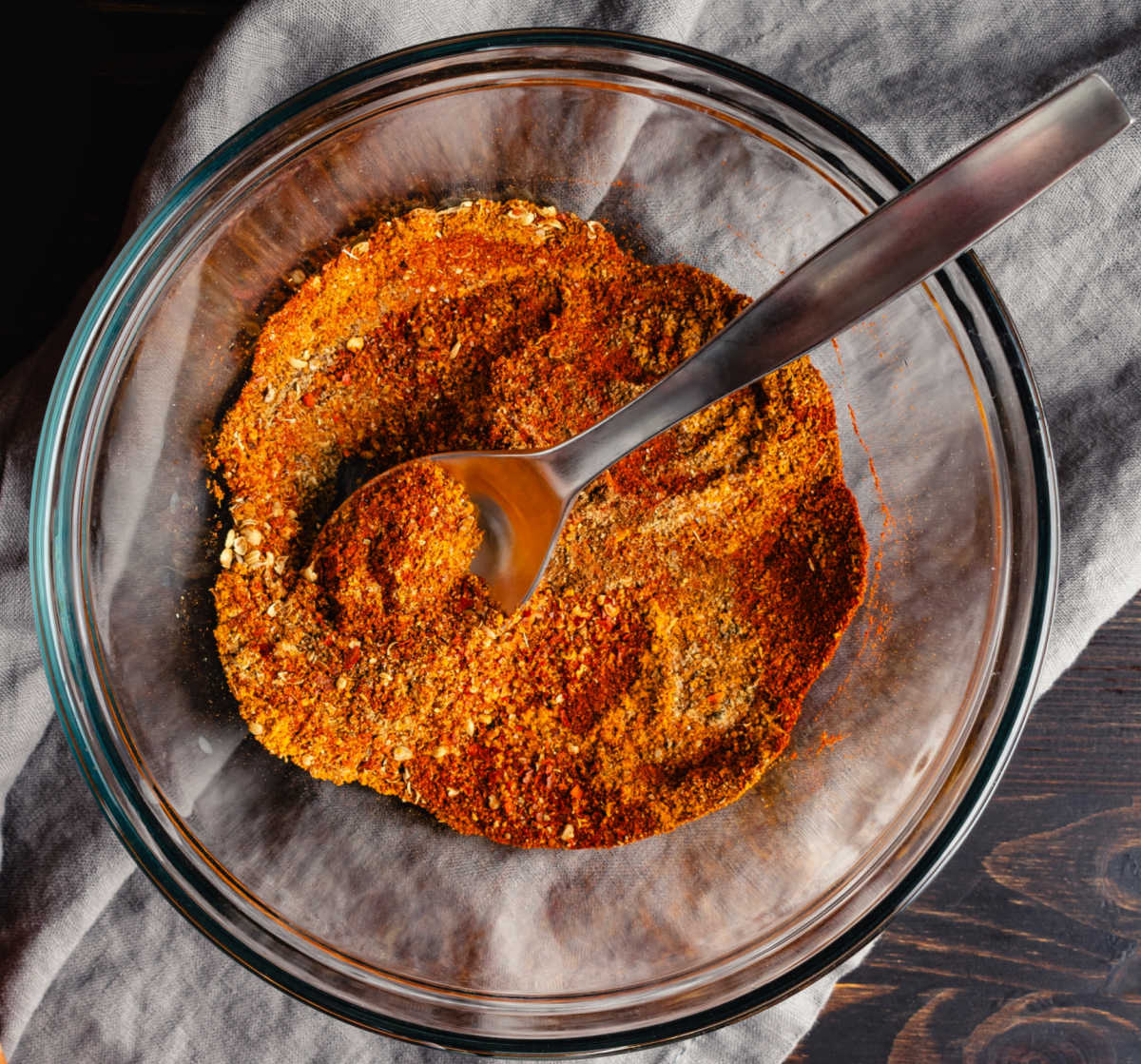Ingredients for Moroccan spice mix in a glass bowl with a spoon on a white tea towel.
