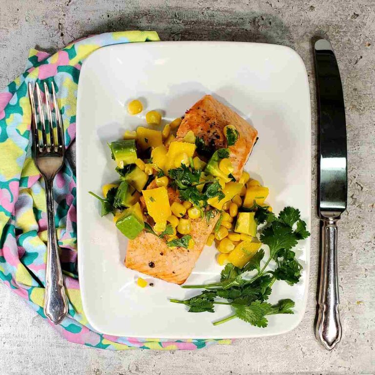 Salmon on a white plate with mango corn salsa and cilantro. Cutlery and napking neawby.