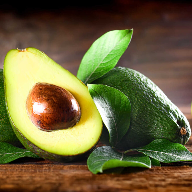 Avocado half and whole fruit with leaves on a wooden counter.
