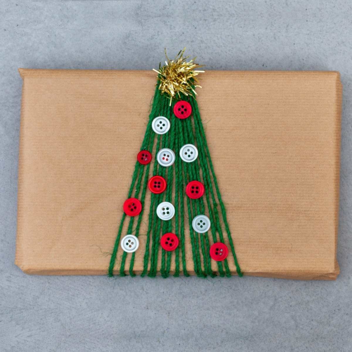 A creative holiday gift wrapping idea showing a rectangular package wrapped in brown paper, with green string wrapped around it to resemble a tree, buttons glued down to stand in for ornaments and a small piece of garland at the top to resemble a star tree topper.