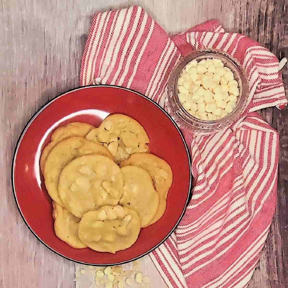 White chocolate macadamia nut cookies A red plate filled with white chocolate macadamia nut cookies with a red and white striped napkin and a bowl of white chocolate chips beside it.
