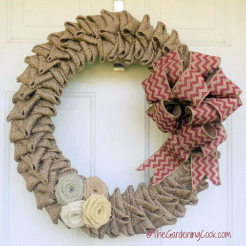 A DIY burlap wreath with a large orange striped bow and cluster of burlap flowers hanging on a front door.
