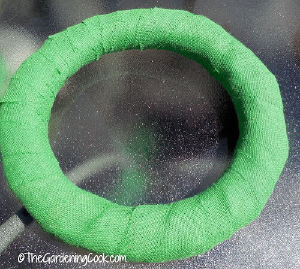 A round wreath form covered in green burlap on a glass table.