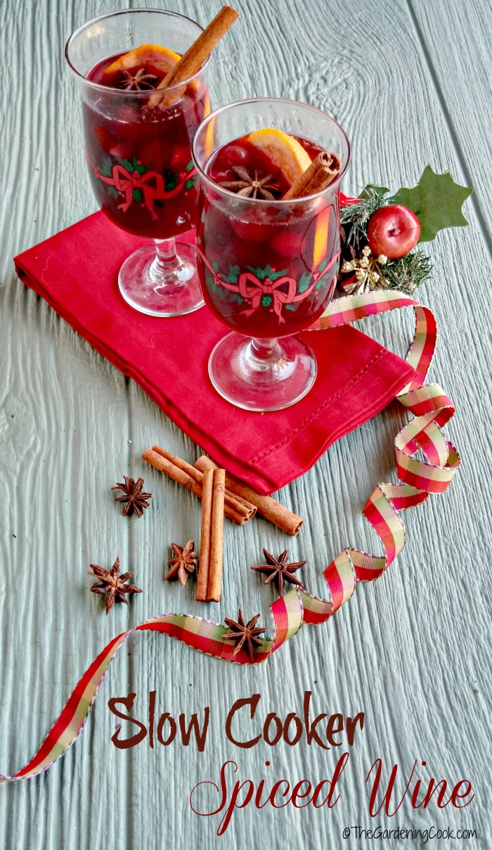 Spiced wine in goblets with lemon slices and cinnamon sticks near some festive items and a red napkin with words reading Slow Cooker Spiced Wine.