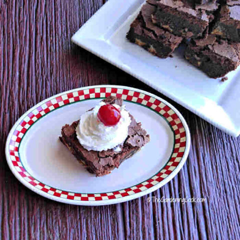 Brownie on a checked plate with whip cream and a cherry, near a plate of brownies.