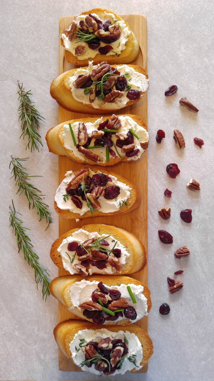 Wooden serving board with Cranberry pecan crostini appetizers on it and cranberry, nuts and rosemary to the sides.