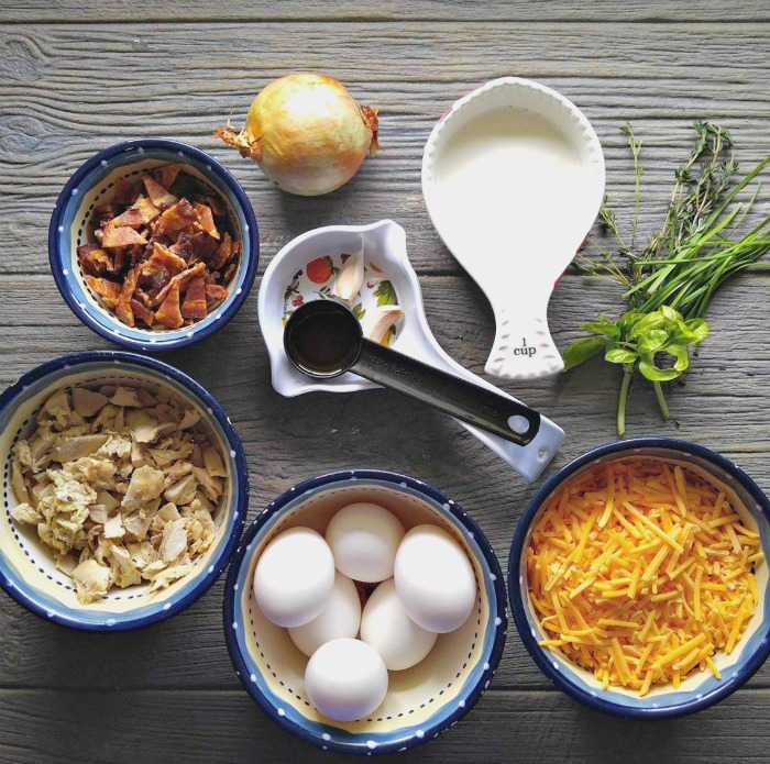 Bacon, onion, garlic, chives, basil, thyme, cheddar cheese, heavy cream, eggs and chicken on a wooden countertop.