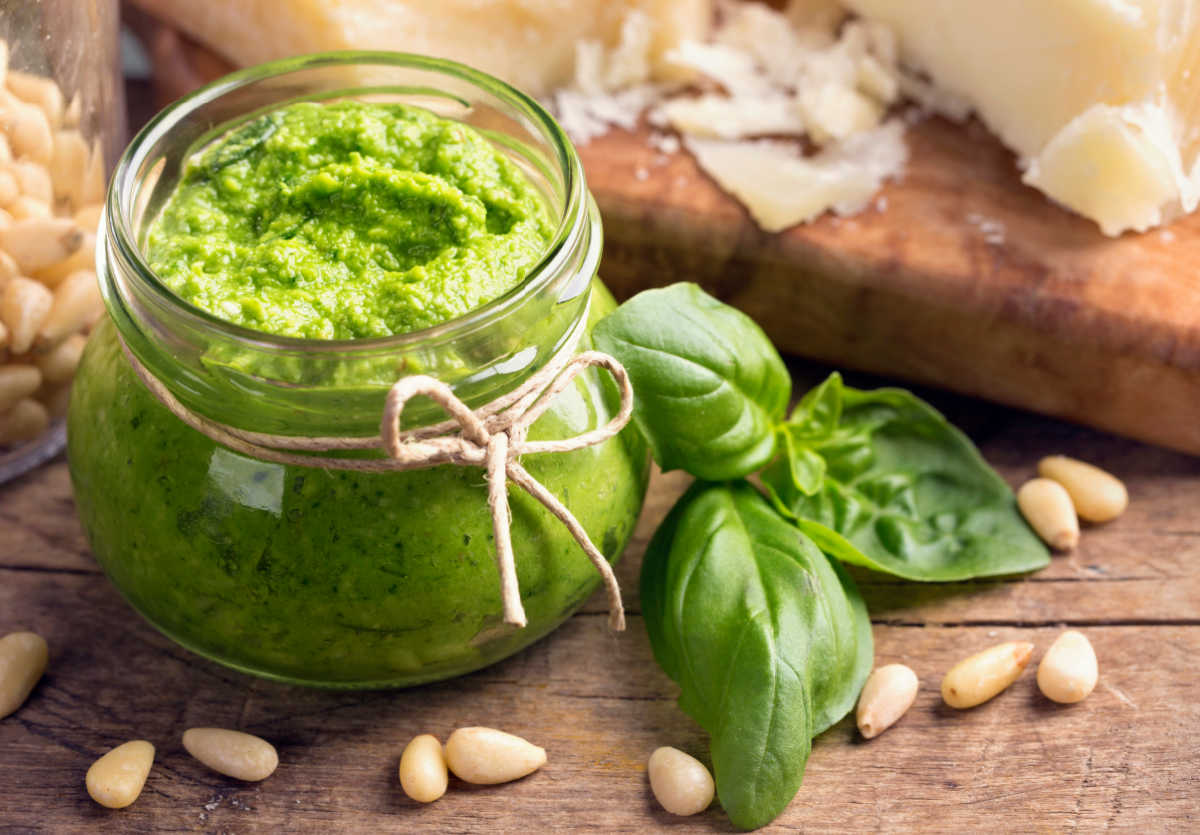 Basil pesto in a glass jar on a wooden counter next to fresh basil, pine nuts and parmesan cheese.