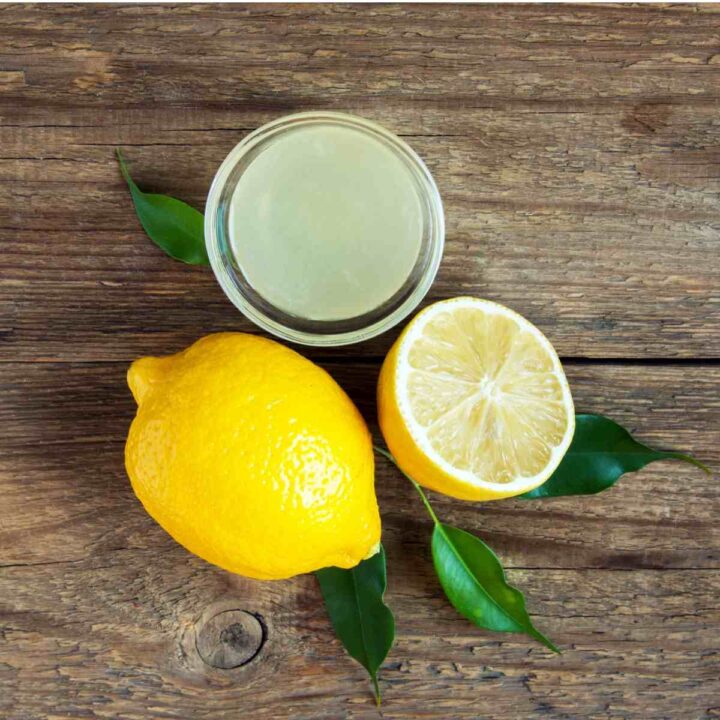 A glass ramekin of freshly squeezed lemon juice above a whole lemon, and halved lemon with several lemon leaves around them.