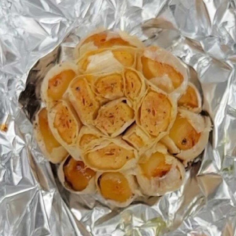 A head of roasted garlic with the tops of the cloves trimmed off sitting in aluminum foil, which has just been cooked following a roasted garlic recipe.