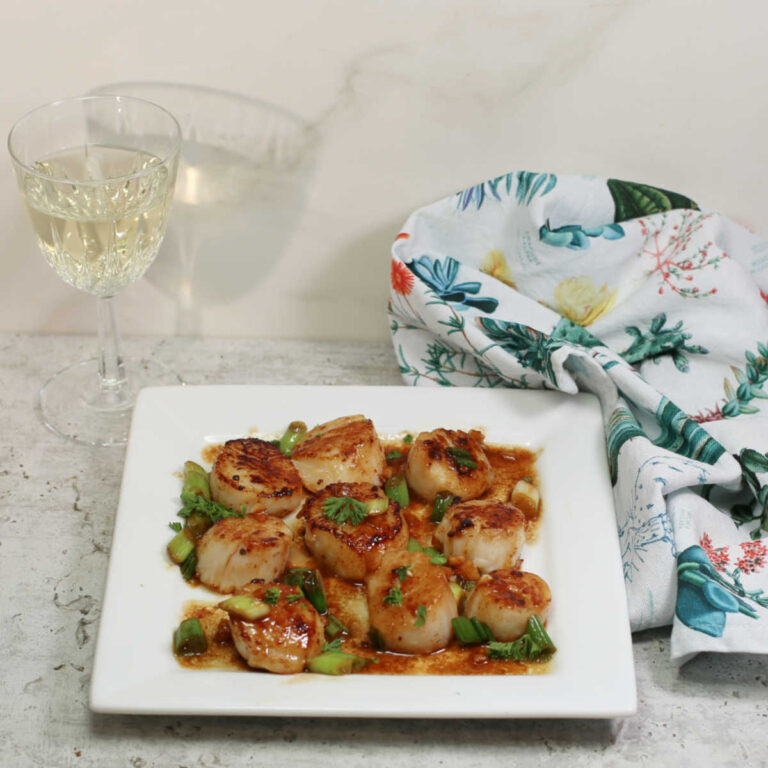 Scallops with white wine butter sauce on a white plate with a glass of white wine and folded napkin.