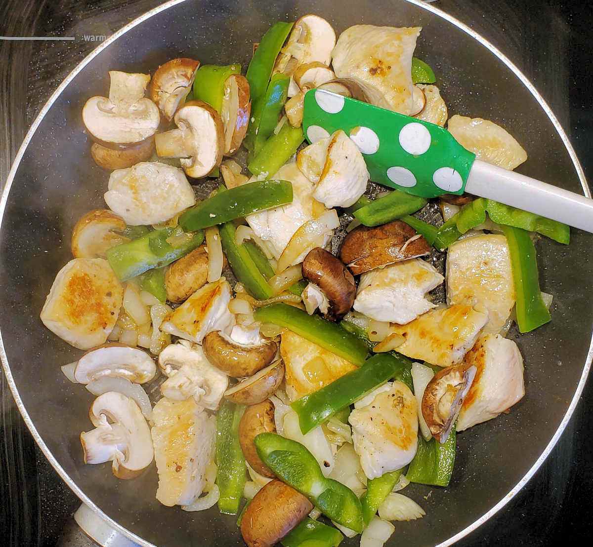 A non-stick pan with browned chicken pieces, and cooked vegetables near a green dotted spatula.