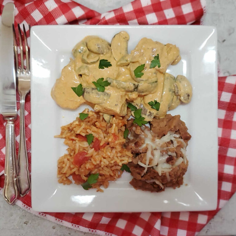 Pollo a la crema with Mexican rice and refried beans on a white plate with red checked napkin.