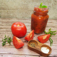 A jar of tomato sauce next to a beefsteak tomato, tomato slices, fresh herbs and a wooden container of salt and pepper.