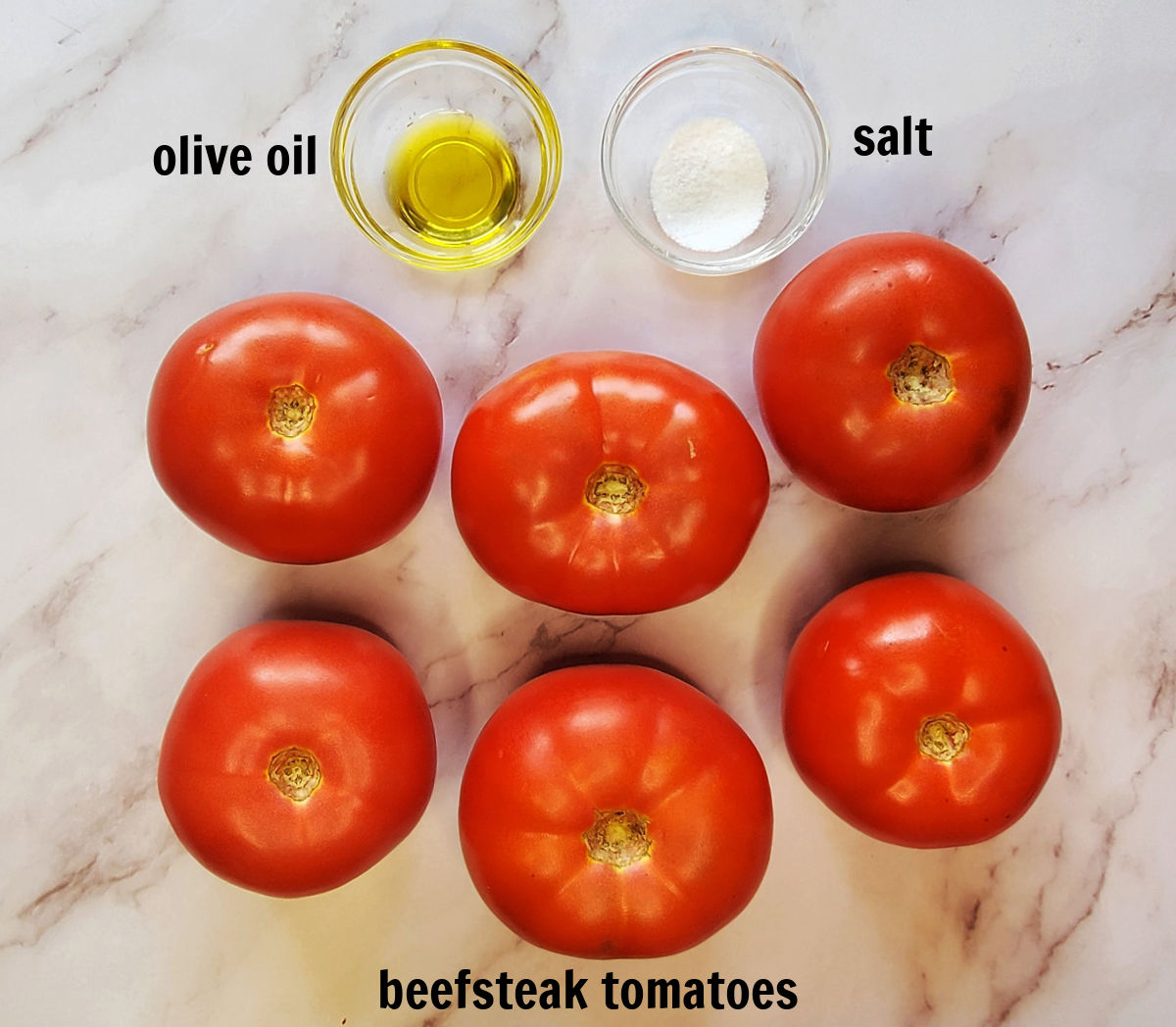 6 beefsteak tomatoes, and two bowls with olive oil and salt, labeled with black text, on a marble countertop - ingredients for roasting tomatoes for sauce.
