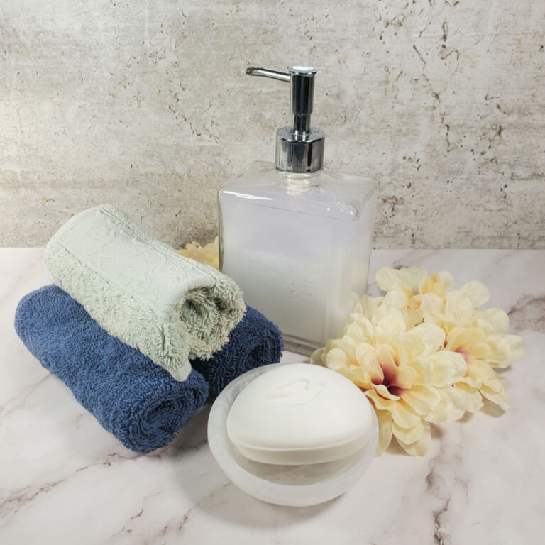 A jar of liquid soap, a bar of soap, flowrs and rolled towels on a counter.