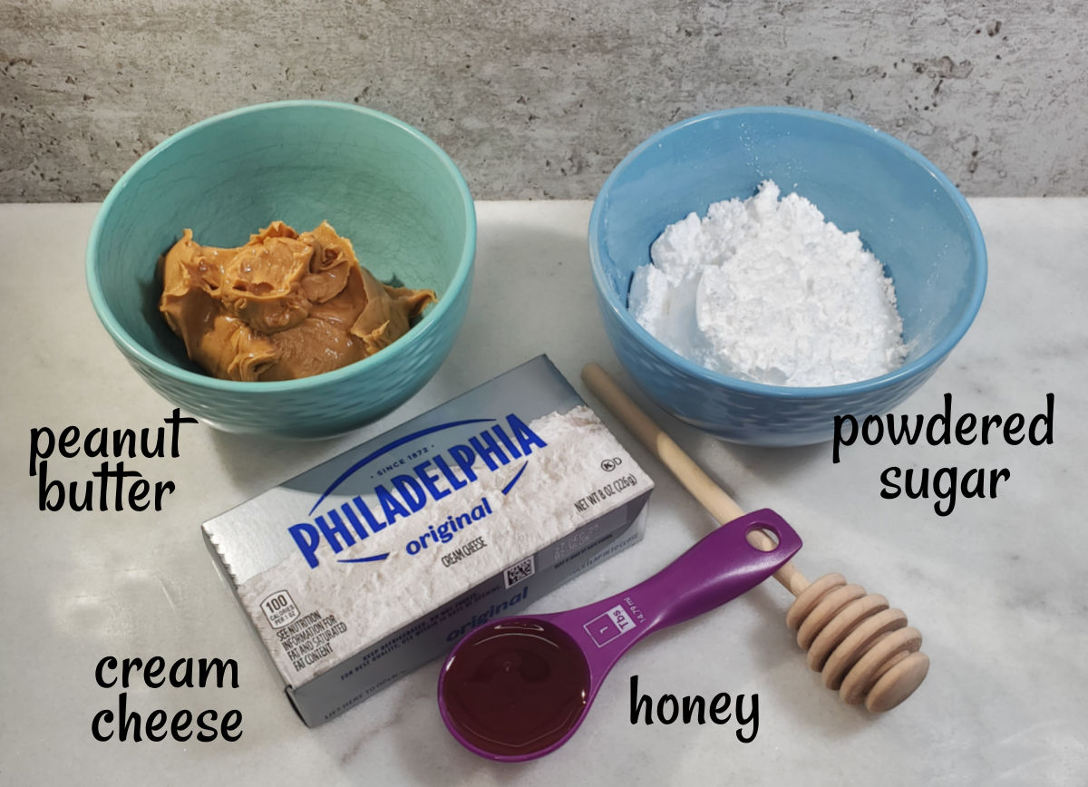 Ingredients for the peanut butter cream cheese frosting Bowls of peanut butter and powdered sugar, next to a package of cream cheese, and spoon of honey resting on a honey dipper.