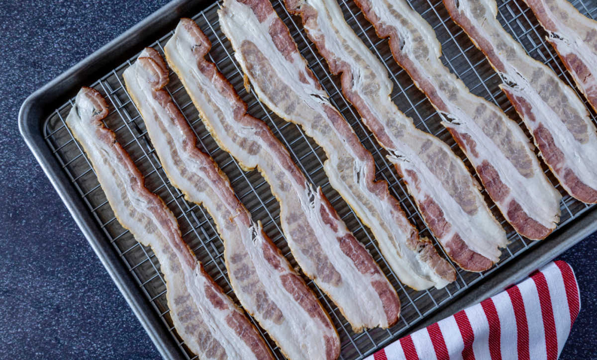 Bacon slices on a wire rack over a baking sheet next to a red and white towel.