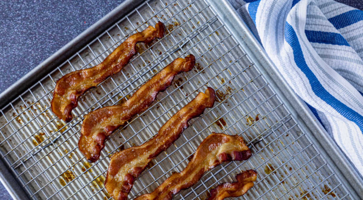 Slices of crispy bacon cooked in the oven on a wire rack and baking pan next to a blue striped towel.