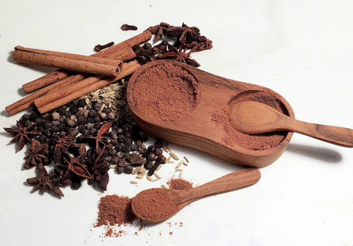 Chinese five spice powder recipe in a wooden dispenser with spoons and whole spices.