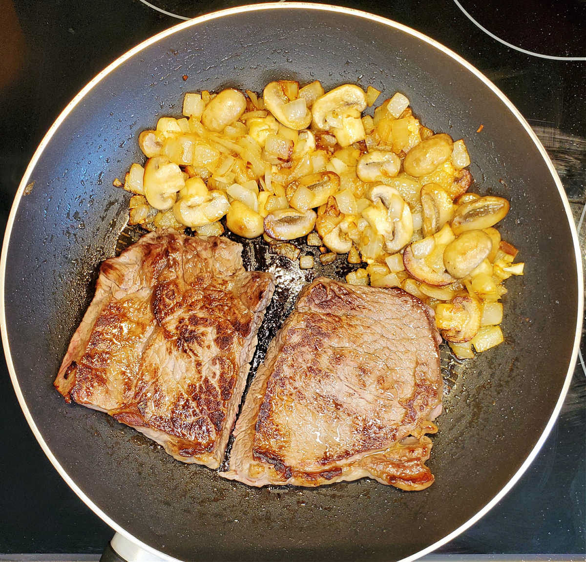 Seared sirloin steak in a skillet next to some cooked mushrooms, garlic and onions.